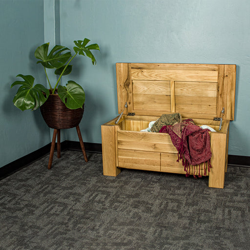 An overall view of the Camden Oak Blanket Box, which is open with blankets hanging out of it. There is a free standing potted plant next to it.
