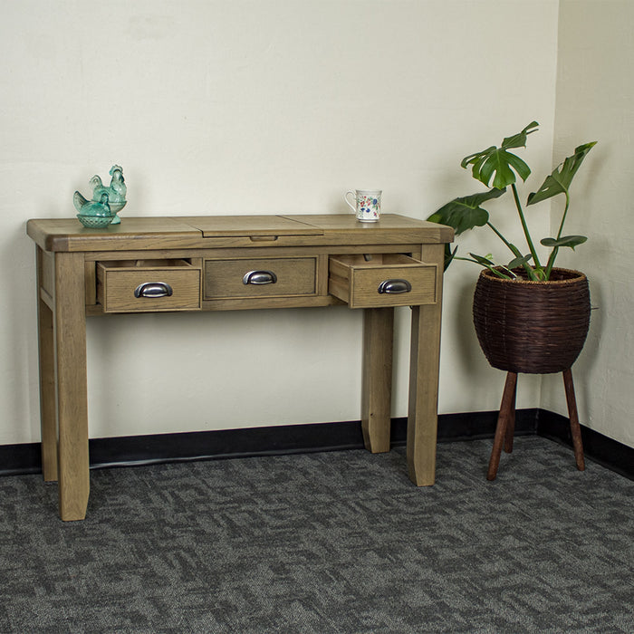 The front of the Houston Oak Dressing Table With Mirror with its drawers open. There are two blue glass ornaments on top to the left and a coffee mug on top to the right. There is a free standing potted plant to the right of the desk.