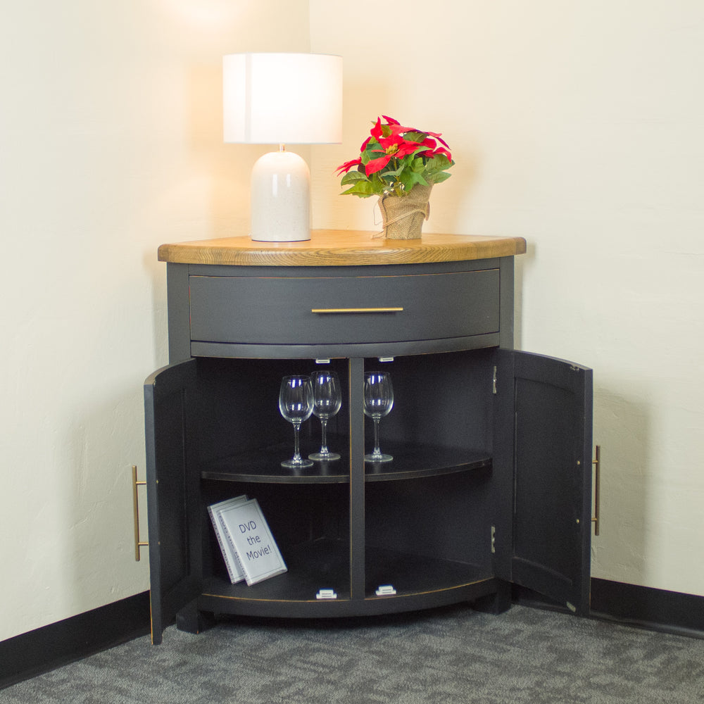 The front of the Cascais Oak-Top Corner Cabinet. There is a lamp and a pot of red flowers on top. There are three wine glasses on the top shelf and two DVDs on the bottom shelf.